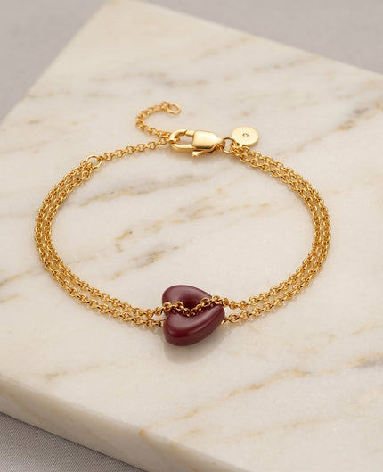 A gold chain bracelet with a small, smooth, dark red heart-shaped charm, displayed on a white marble slab.
