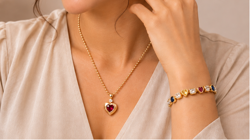 A woman wearing a gold heart pendant necklace with a red heart-shaped stone, a matching gold heart bracelet, and a purple teardrop earring, shown in a warm, softly lit setting.